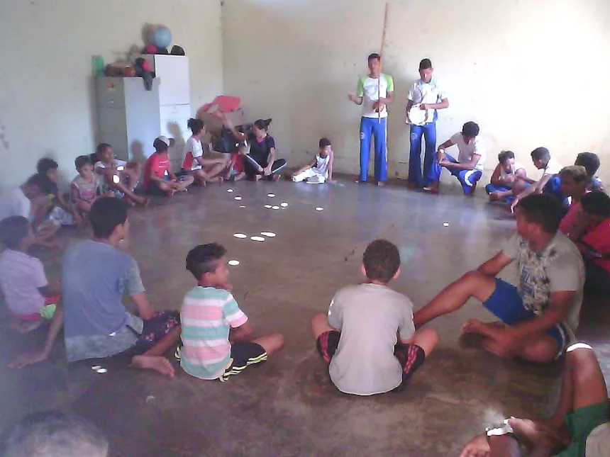 Roda de capoeira com crianças e adolescentes sentados no chão do salão comunitário